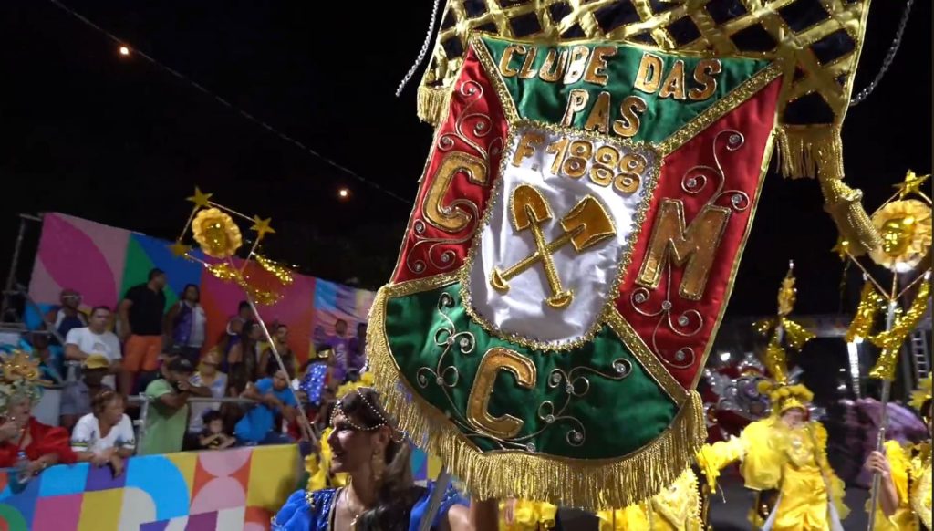A cena mostra um desfile carnavalesco cheio de cores e detalhes. No centro, há um grande estandarte ornamentado com franjas douradas e símbolos de pás cruzadas, trazendo inscrições que remetem a um clube fundado em 1888. Ao redor do estandarte, pessoas vestem fantasias elaboradas, em tons vivos de amarelo, com enfeites de estrelas e flores nas cabeças. Ao fundo, espectadores assistem sentados diante de uma parede multicolorida.