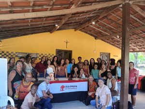 Um grupo de pessoas está reunido em um espaço coberto, posando para foto em torno de uma mesa com bolo. Sobre a mesa há uma toalha escrita ‘Quilombos de Pernambuco’, com um símbolo de árvore vermelha.