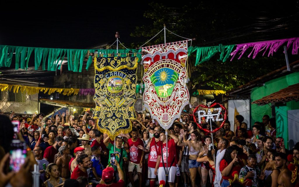 Essa imagem mostra uma festa popular realizada à noite em uma rua cheia de pessoas. A multidão veste roupas festivas, muitas em vermelho, e duas bandeiras grandes e ornamentadas se destacam no centro. Uma delas traz o nome “Cariri” com bordados em azul e dourado; a outra celebra os 70 anos do grupo “Elefante de Olinda”, com bordados coloridos e a figura de um elefante sob o sol. Acima da rua, há fitas verdes e amarelas penduradas, e também um letreiro em forma de coração com a palavra “Paz”. O clima é alegre, vibrante e transmite a energia de uma celebração cultural tradicional brasileira.