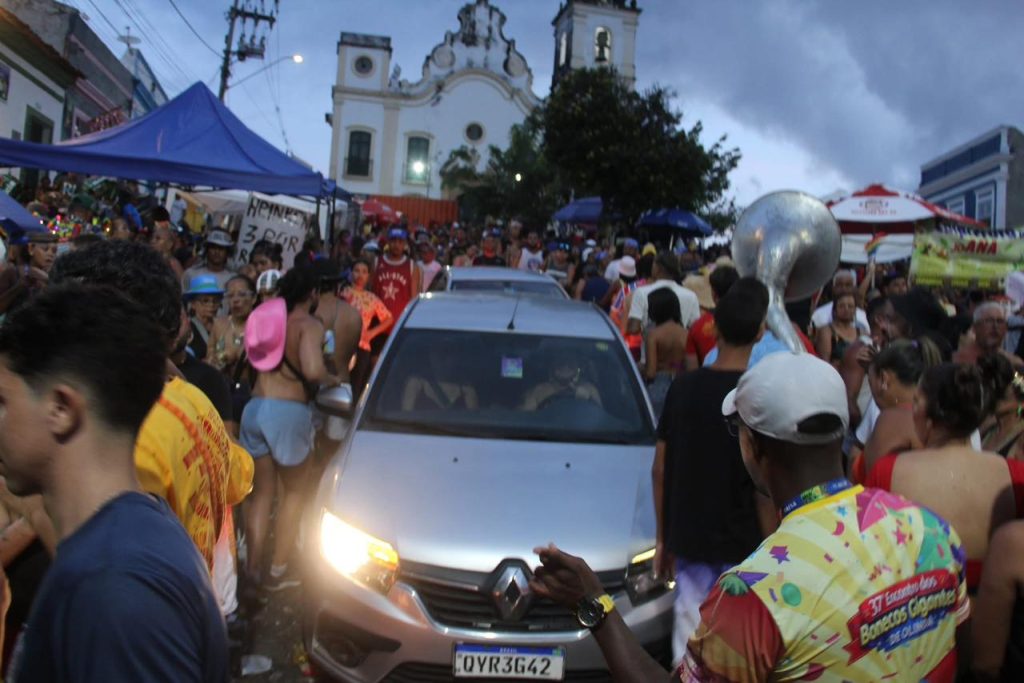 A foto mostra uma rua cheia de pessoas participando de uma celebração popular. Há barracas montadas dos dois lados, gente fantasiada e até um músico com um grande instrumento de sopro, o que sugere um clima de desfile ou carnaval. Um carro prateado passa devagar pelo meio da multidão, enquanto ao fundo se vê uma igreja histórica com duas torres. O céu nublado dá um tom mais dramático à cena festiva.