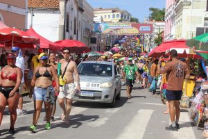 A foto mostra uma rua histórica tomada por foliões durante uma festa popular. As pessoas caminham em clima de celebração, muitas com roupas coloridas e fantasias. De ambos os lados da rua há barracas cobertas por guarda-sóis vermelhos, formando um corredor animado. No primeiro plano, um carro prateado avança devagar entre a multidão. Ao fundo, um grande banner atravessa a rua com mensagens sobre segurança e controle de acesso. As construções coloniais completam o cenário festivo e cultural.