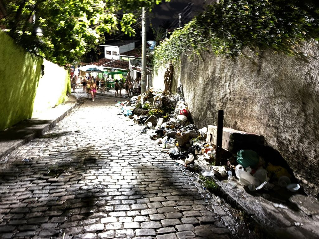 A foto mostra uma rua estreita de paralelepípedos à noite, iluminada por postes de luz. À direita, há uma grande quantidade de lixo acumulado na calçada, com caixas, plásticos e outros objetos descartados junto a um muro coberto de plantas. Do lado esquerdo, aparece uma parede pintada de verde. Mais ao fundo, um grupo de pessoas se reúne perto de barracas ou quiosques sob guarda-sóis, criando um contraste entre o ambiente festivo ao longe e a cena de descuido urbano em primeiro plano.