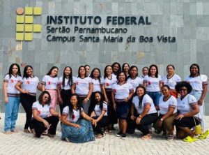 Na foto, há um grupo de jovens mulheres negras reunidas em frente a uma parede com uma placa que identifica o local como o Instituto Federal do Sertão Pernambucano – Campus Santa Maria da Boa Vista. Todas estão vestindo camisetas brancas iguais, com detalhes em vermelho e preto, o que sugere que participam de um evento ou atividade conjunta da instituição. Elas estão lado a lado, sorrindo e posando para a foto.