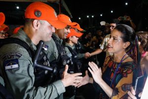 A foto mostra a governadora Raquel Lyra em um evento noturno, cumprimentando policiais militares. Ela está de frente para um dos policiais, apertando a mão dele em gesto de cordialidade. Os policiais usam uniforme escuro com boné laranja e distintivos que indicam patrulhamento tático. Ao redor, há outras pessoas observando e registrando o momento com celulares. O ambiente transmite clima de celebração.