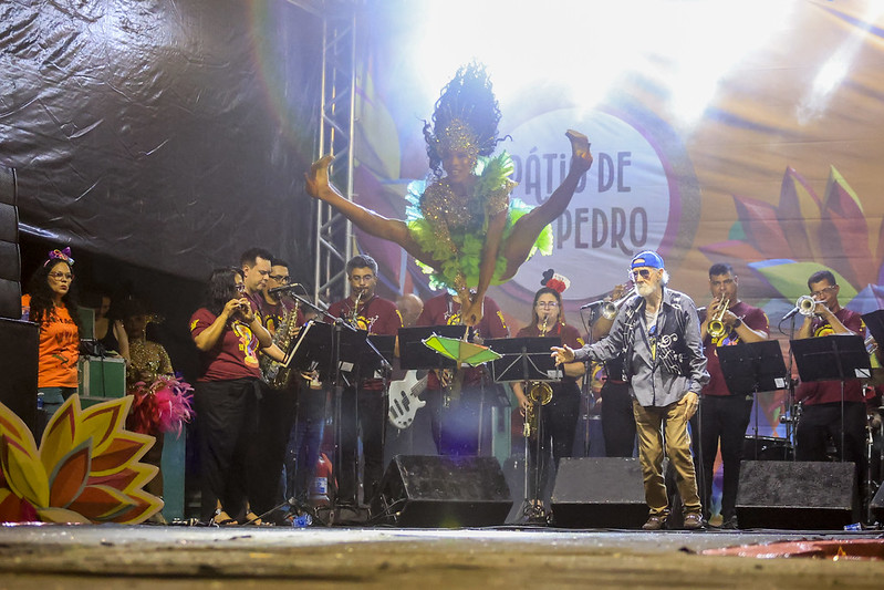 Foto de um palco durante uma apresentação musical ao vivo. Ao centro, uma passista de frevo em pleno salto no ar, com fantasia verde brilhante e grande adorno de cabeça, pernas abertas e braços erguidos. Ao fundo, uma banda com vários músicos tocando instrumentos de sopro diante de estantes de partitura. À direita, um cantor idoso usa boné azul e segura o microfone enquanto se inclina para o público. Luzes fortes iluminam a cena e um painel colorido com a inscrição “Pátio de São Pedro” aparece ao fundo.