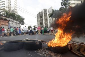 A imagem mostra uma cena de protesto em uma rua de cidade. No primeiro plano, há pneus e pedaços de madeira em chamas formando uma barricada, de onde sai bastante fumaça preta e espessa. Atrás dessa barreira, um grupo de pessoas está reunido, algumas próximas ao fogo, outras observando. O ambiente é claramente urbano: há prédios altos de apartamentos ao fundo, fios elétricos visíveis e alguns veículos estacionados. A mistura de construções modernas e mais antigas compõe o cenário.