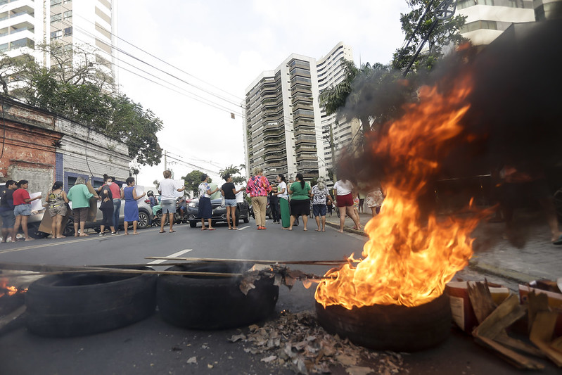 A imagem mostra uma cena de protesto em uma rua de cidade. No primeiro plano, há pneus e pedaços de madeira em chamas formando uma barricada, de onde sai bastante fumaça preta e espessa. Atrás dessa barreira, um grupo de pessoas está reunido, algumas próximas ao fogo, outras observando. O ambiente é claramente urbano: há prédios altos de apartamentos ao fundo, fios elétricos visíveis e alguns veículos estacionados. A mistura de construções modernas e mais antigas compõe o cenário.