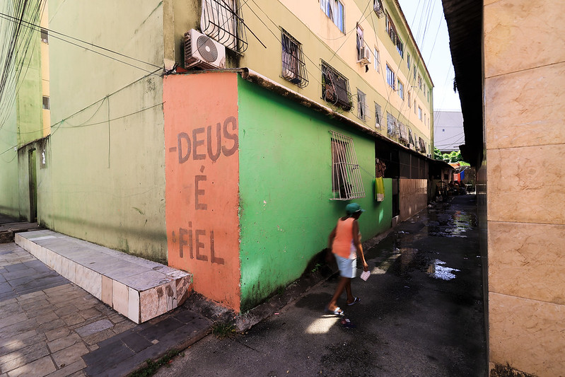 A imagem mostra um beco estreito entre dois prédios. O prédio à esquerda tem paredes em verde e bege, janelas com grades e um aparelho de ar-condicionado instalado na parte superior. Em um canto, há uma faixa vertical vermelha com letras grandes que dizem “DEUS É FIEL”. O chão é pavimentado e apresenta algumas áreas molhadas que refletem a luz. No centro da cena, uma pessoa caminha pelo beco. Ela veste boné verde, camiseta laranja e shorts azuis, segurando um pequeno objeto em uma das mãos. Ao fundo, o beco se prolonga, revelando mais construções e um pouco de vegetação.