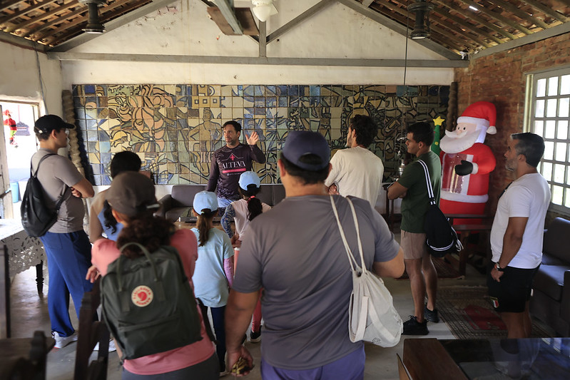 Um grupo de pessoas, incluindo crianças, escuta um homem diante de um mural colorido em uma sala rústica. Há paredes de tijolos, vigas de madeira e um grande Papai Noel inflável ao lado.