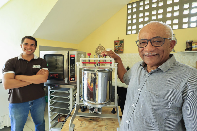 Dois homens estão sorrindo em uma cozinha profissional. Um homem idoso segura um equipamento de aço inox, enquanto o outro, bem mais jovem, está um pouco a frente de um forno grande com bandejas e paredes amarelas decoradas.