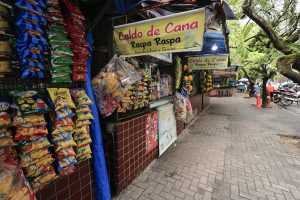 A imagem mostra uma cena típica de comércio de rua. Há várias barracas pequenas alinhadas ao longo de uma calçada pavimentada, cobertas por toldos coloridos. Nas barracas, penduram-se pacotes de salgadinhos e outros produtos, enquanto algumas oferecem bebidas e comidas frescas. Uma placa visível anuncia “Caldo de Cana, Rasp Rasp, Salgados e Sucos”, indicando que ali se vendem caldo de cana, raspadinha e lanches. As árvores grandes ao redor fornecem sombra, criando um ambiente agradável para os pedestres que circulam. Também se veem motocicletas estacionadas na rua, reforçando o clima movimentado e cotidiano do local.