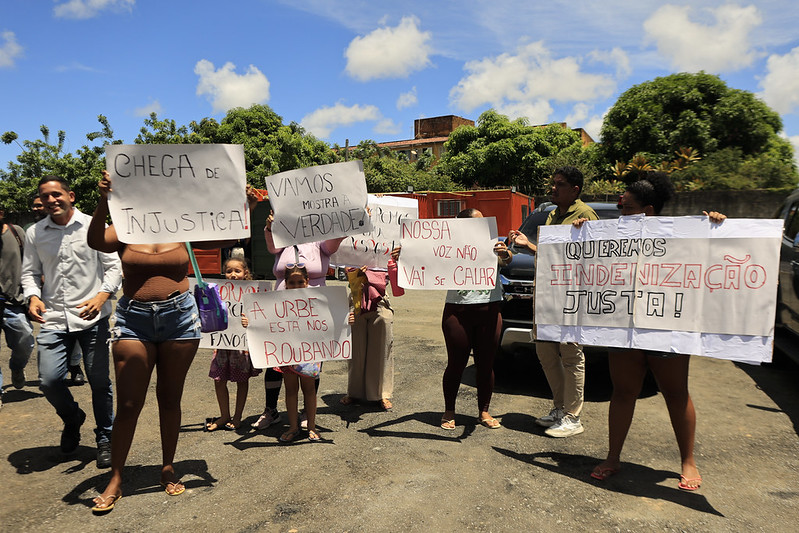 A imagem mostra um grupo de pessoas reunidas em um espaço aberto, em um dia ensolarado. Elas estão segurando cartazes de protesto escritos em português, expressando indignação e exigindo justiça. Entre as mensagens visíveis estão frases como: “CHEGA DE INJUSTIÇA!”, “VAMOS MOSTRAR A VERDADE”, “NOSSA VOZ NÃO VAI SE CALAR”, “A URB ESTÁ NOS ROUBANDO”, “QUEREMOS INDENIZAÇÃO JUSTA!” O cenário ao redor inclui árvores, uma cerca vermelha e alguns prédios ao fundo. A atmosfera transmitida é de mobilização social.