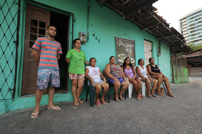 A foto mostra várias pessoas sentadas em cadeiras em frente a uma casa simples, com parede pintada de verde