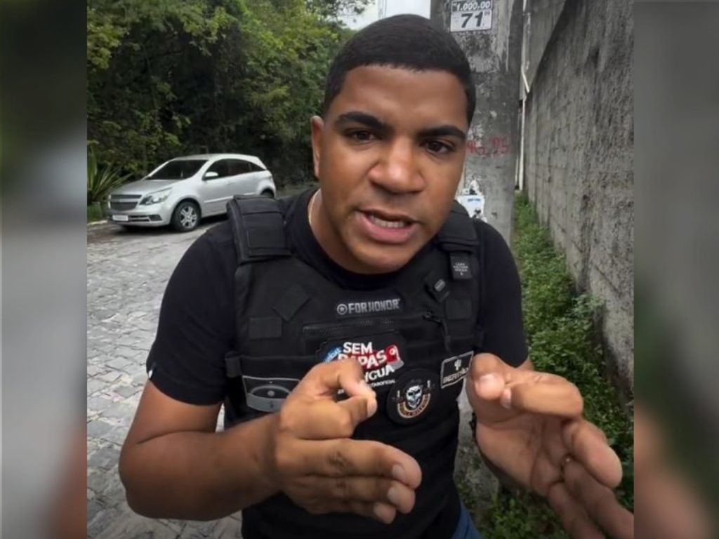 A foto mostra Emerson Jhuan, um homem negro de pele clara, cabelos curtos e escuros e barba rala, em pé sobre uma rua de paralelepípedos ao lado de um muro de concreto com plantas verdes; ele veste um colete tático preto com vários patches, além de um distintivo com a palavra “DEPARTAMENTO”; Emerson aparece gesticulando com a mão em direção à câmera, transmitindo energia e expressividade, enquanto ao fundo se vê um carro prateado estacionado e um poste com um cartaz de números.