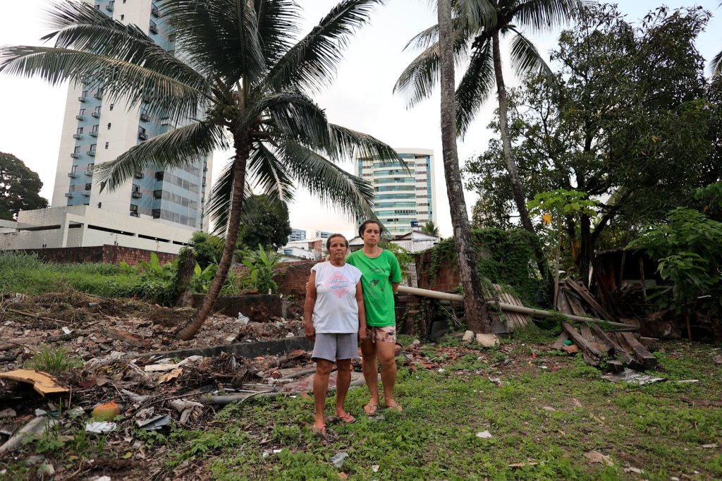 A foto mostra duas pessoas em pé sobre um terreno irregular, coberto por entulho e pedaços de madeira. Ao fundo, há palmeiras altas e vegetação densa, contrastando com prédios modernos e bem cuidados que aparecem atrás. A cena evidencia o contraste entre um espaço degradado em primeiro plano e a urbanização organizada ao fundo.