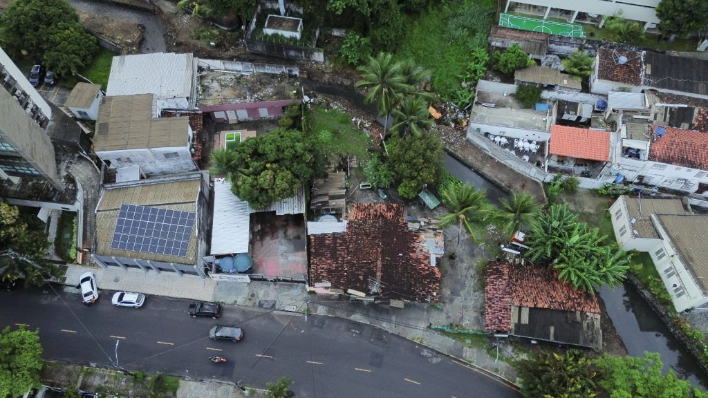 A foto mostra uma vista aérea de um bairro urbano com construções variadas — algumas casas com telhados de telha, outras com coberturas de metal. Um dos prédios tem painéis solares instalados no telhado. Na parte inferior, há uma rua pavimentada com carros estacionados e em movimento. Um canal estreito atravessa a área, ladeado por vegetação e palmeiras. Mais ao fundo, aparecem outros edifícios e uma pequena quadra esportiva verde.