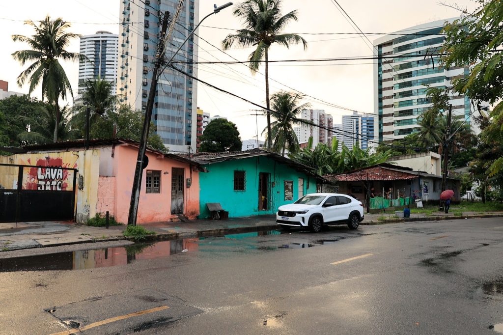 A foto mostra uma rua molhada com poças d’água, sugerindo que choveu recentemente. No primeiro plano, há duas casas simples — uma pintada de pêssego com a placa “LAVA JATO” e outra em tom turquesa. Um carro branco está estacionado ao lado. Ao fundo, aparecem prédios altos e modernos, além de palmeiras e fios elétricos, criando um contraste entre construções modestas e a urbanização verticalizada da região.