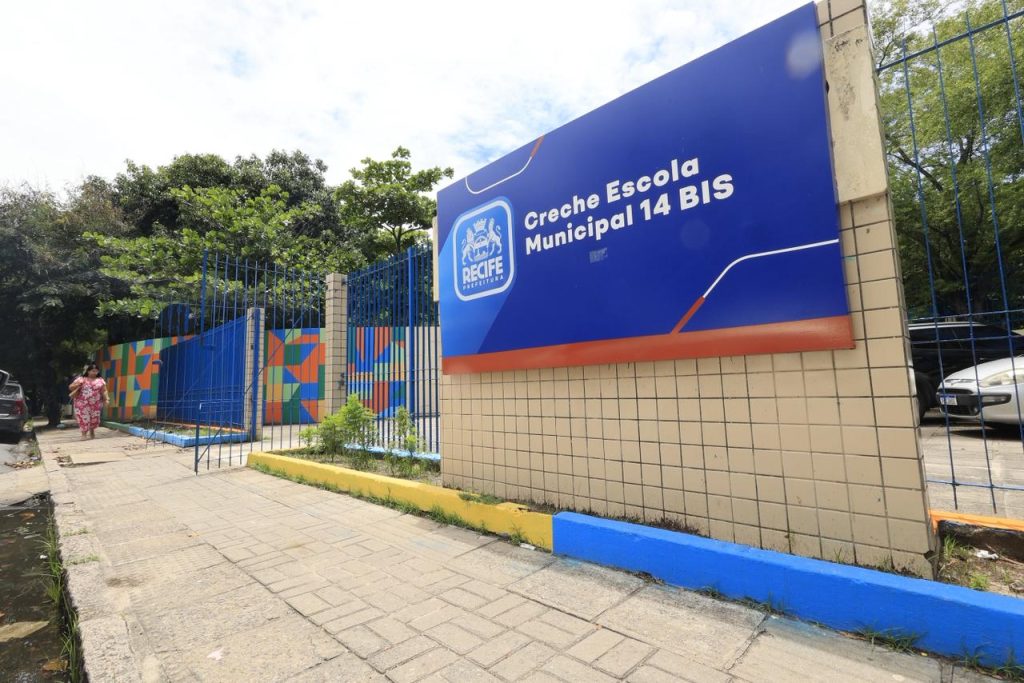 A entrada mostra um letreiro escrito “Creche Escola Municipal 14 BIS”. Abaixo está o símbolo da Prefeitura do Recife. O muro tem desenhos geométricos coloridos em azul, laranja, verde e amarelo. Há árvores próximas, uma calçada e uma pessoa caminhando.