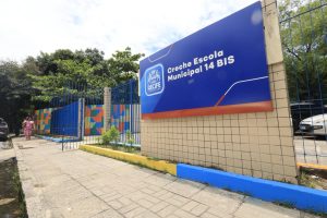 A entrada mostra um letreiro escrito “Creche Escola Municipal 14 BIS”. Abaixo está o símbolo da Prefeitura do Recife. O muro tem desenhos geométricos coloridos em azul, laranja, verde e amarelo. Há árvores próximas, uma calçada e uma pessoa caminhando.