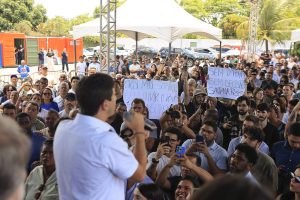 A foto mostra uma manifestação pública ao ar livre. Há uma multidão reunida sob tendas, ouvindo Joã0o Campos falando ao microfone. Muitas pessoas estão gravando ou tirando fotos com seus celulares. Entre os cartazes erguidos, alguns trazem mensagens de protesto, como críticas a decisões tomadas sem ouvir a população e frases de resistência ligadas ao bairro Santana. É uma cena de engajamento coletivo, com forte participação popular.