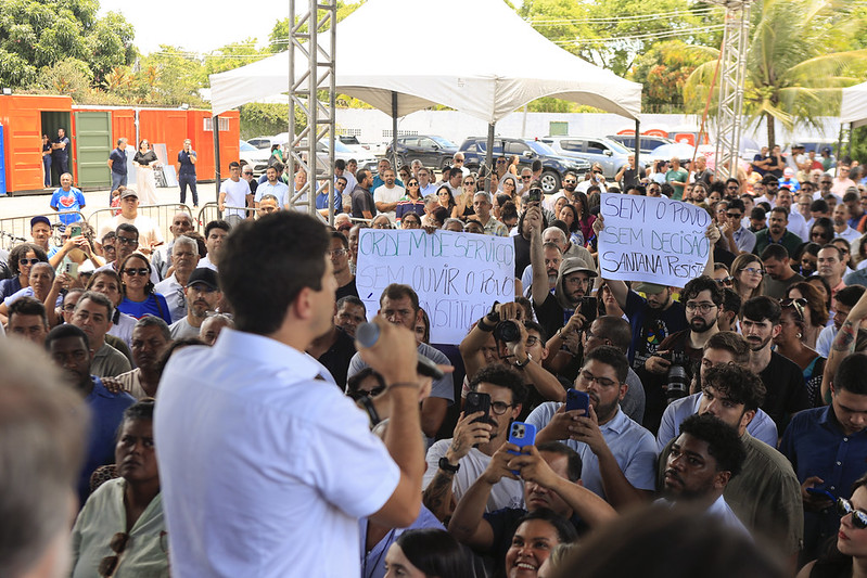A foto mostra uma manifestação pública ao ar livre. Há uma multidão reunida sob tendas, ouvindo Joã0o Campos falando ao microfone. Muitas pessoas estão gravando ou tirando fotos com seus celulares. Entre os cartazes erguidos, alguns trazem mensagens de protesto, como críticas a decisões tomadas sem ouvir a população e frases de resistência ligadas ao bairro Santana. É uma cena de engajamento coletivo, com forte participação popular.