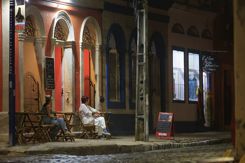 A foto mostra uma cena noturna em uma rua de paralelepípedos em Olinda, com atmosfera tranquila e acolhedora. Duas pessoas estão sentadas em cadeiras de madeira diante de um prédio colorido, com arcos e grades de ferro trabalhadas. Há placas e letreiros que indicam comércio local, incluindo um bar com uma lousa anunciando os preços das bebidas e uma loja de moda alternativa. As fachadas em vermelho, laranja e azul, junto com a iluminação suave, criam um ambiente culturalmente rico e convidativo.