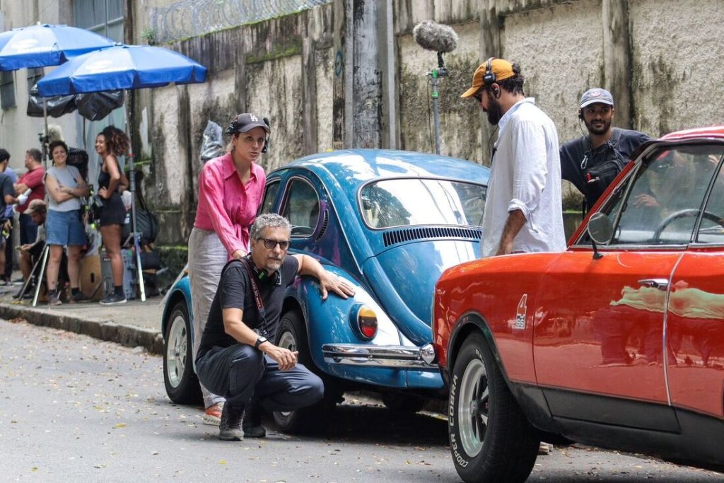 A foto mostra uma equipe de filmagem trabalhando em uma rua. Em primeiro plano, aparecem dois carros antigos: um Fusca azul e outro carro vermelho com pneus marcados com “Cooper Cobra”. Ao redor deles, várias pessoas da produção estão em ação — um homem agachado ao lado do Fusca, uma mulher de camisa rosa e calça branca, e outro homem com fones de ouvido. Acima, vê-se um microfone de boom captando o som. Mais ao fundo, há equipamentos e membros da equipe sob guarda-sóis azuis, compondo o ambiente típico de bastidores de um set de filmagem.