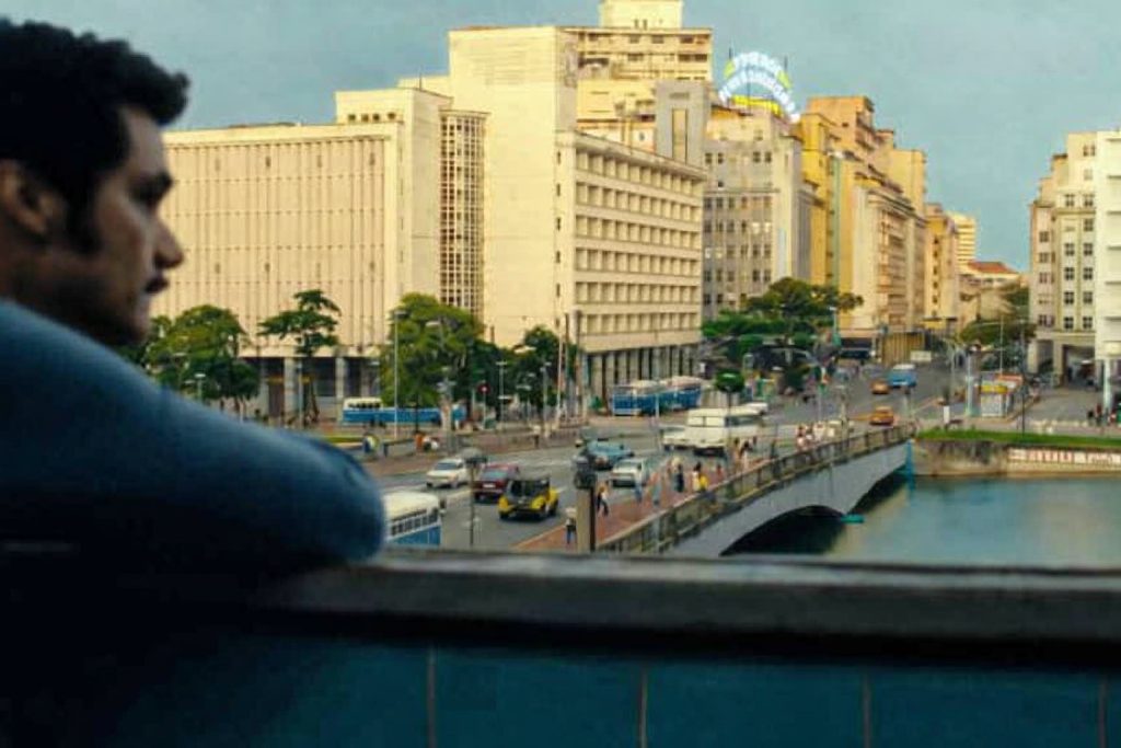 Foto célebre do filme O Agente Secreto em que se vê a silhueta de um homem, o ator Wagner Moura, encostado em uma sacada, observando a movimentação intensa do centro do Recife. À frente, há uma ponte sobre o rio Capibaribe, por onde passam carros, ônibus e pedestres. Ao fundo, aparecem prédios de estilo mais antigo, com algumas placas visíveis, compondo um cenário urbano vibrante.