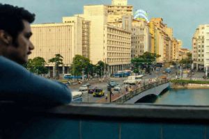 Foto célebre do filme O Agente Secreto em que se vê a silhueta de um homem, o ator Wagner Moura, encostado em uma sacada, observando a movimentação intensa do centro do Recife. À frente, há uma ponte sobre o rio Capibaribe, por onde passam carros, ônibus e pedestres. Ao fundo, aparecem prédios de estilo mais antigo, com algumas placas visíveis, compondo um cenário urbano vibrante.