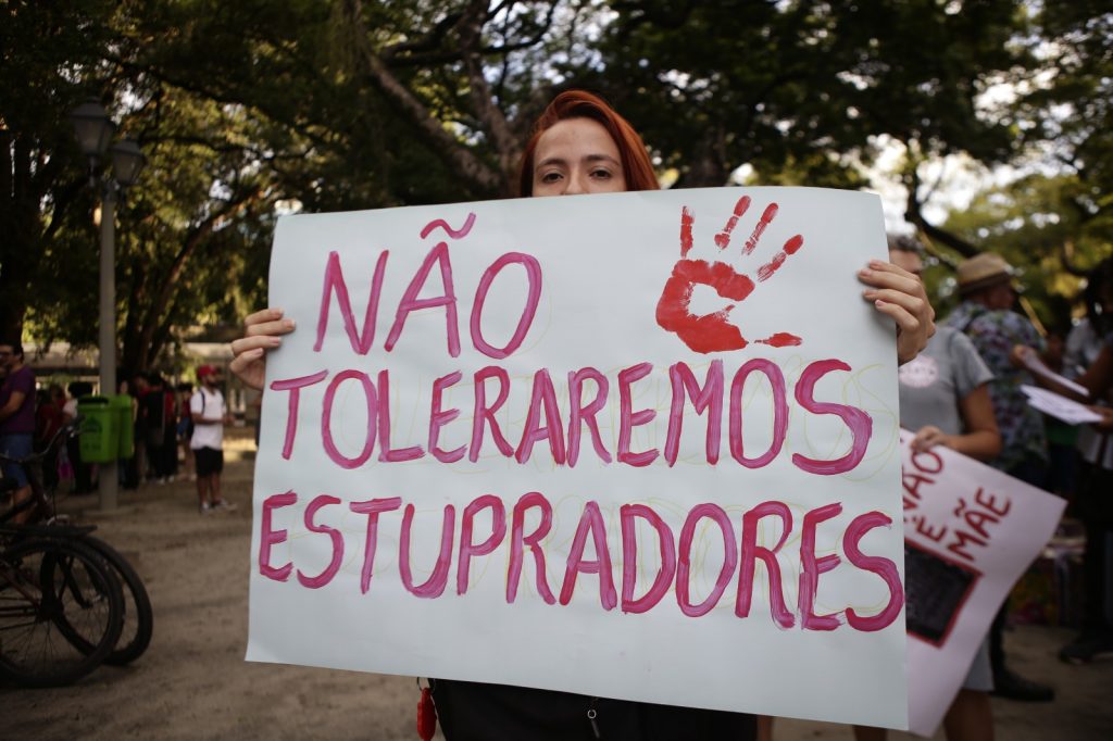 A foto mostra uma cena de manifestação em espaço público. No centro, uma pessoa segura um cartaz branco com letras grandes em português que dizem “NÃO TOLERAREMOS ESTUPRADORES”, acompanhado de uma marca de mão vermelha. Ao fundo, outras pessoas também participam do protesto. A imagem transmite uma mensagem clara de repúdio à violência sexual e de mobilização coletiva contra abusos.