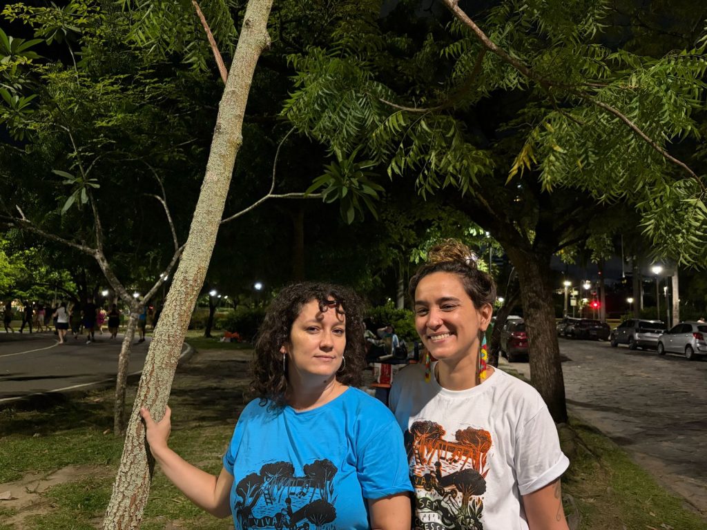 Na foto aparecem duas mulheres em pé, à noite, diante de uma árvore em um espaço arborizado e iluminado por postes de luz. À esquerda está Manuela Galindo, que veste camiseta azul com a palavra “VIVA” e ilustração de árvores. Ela tem pele clara, cabelos castanhos escuros presos e porte médio, segurando o tronco da árvore. À direita está Cecília da Fonte, usando camiseta branca com estampa em laranja e preto também com a palavra “VIVA”. Ela tem pele clara, cabelos longos e escuros soltos, sorriso aberto e porte esguio. Ao fundo, vê-se uma pista com pessoas caminhando ou correndo, além de carros estacionados e mais árvores, compondo um ambiente urbano e verde.