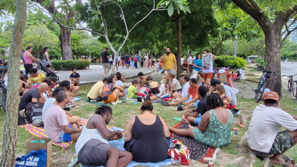 A foto mostra um encontro comunitário ao ar livre, em um parque sombreado por árvores. Muitas pessoas estão sentadas em mantas e esteiras, participando de atividades coletivas como escrever, desenhar e conversar. Há uma mesa com materiais e alguém segurando um cartaz, o que indica que se trata de um evento organizado. O ambiente transmite interação social, criatividade e convivência em grupo.
