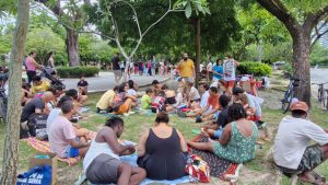 A foto mostra um encontro comunitário ao ar livre, em um parque sombreado por árvores. Muitas pessoas estão sentadas em mantas e esteiras, participando de atividades coletivas como escrever, desenhar e conversar. Há uma mesa com materiais e alguém segurando um cartaz, o que indica que se trata de um evento organizado. O ambiente transmite interação social, criatividade e convivência em grupo.