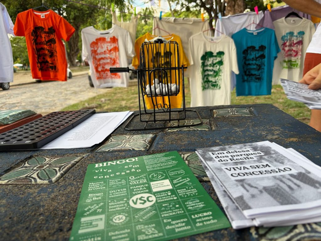 A imagem mostra uma cartela verde de bingo, com várias camisetas ilustradas atrás, em um varal no parque