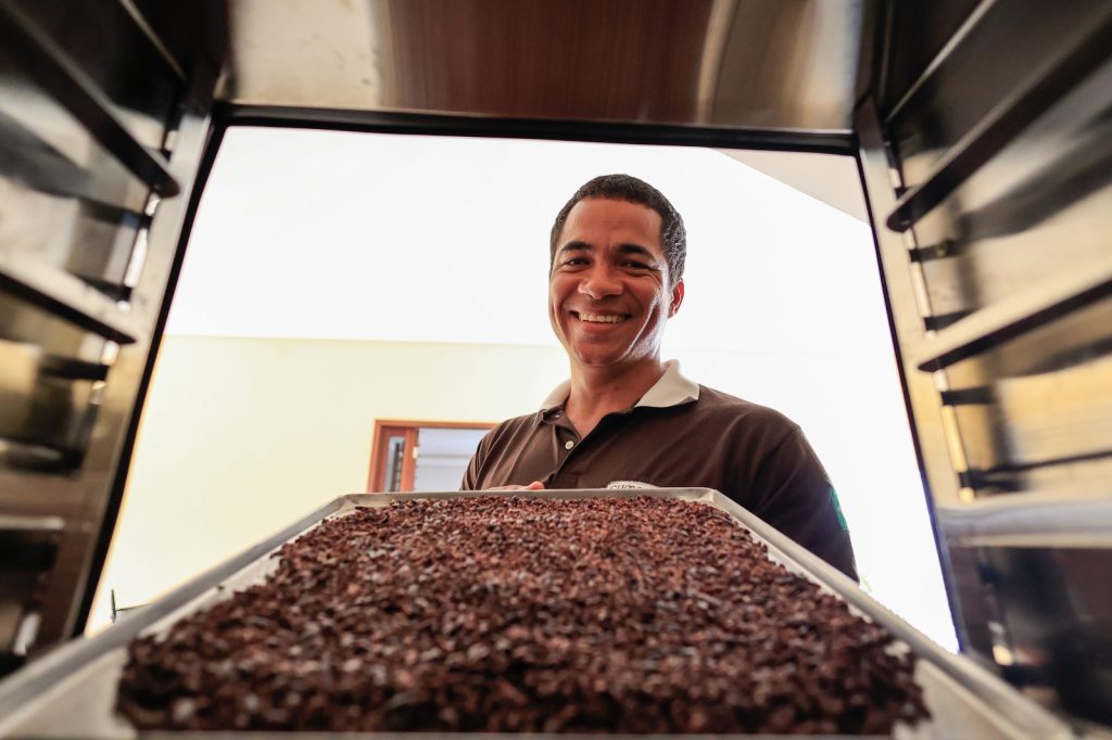 Foto de homem pardo, de cabelos curtos, usando camisa preta de gola polo branca retira de um forno ou de uma armário uma grande bandeja repleta de sementes marrons de cacau.