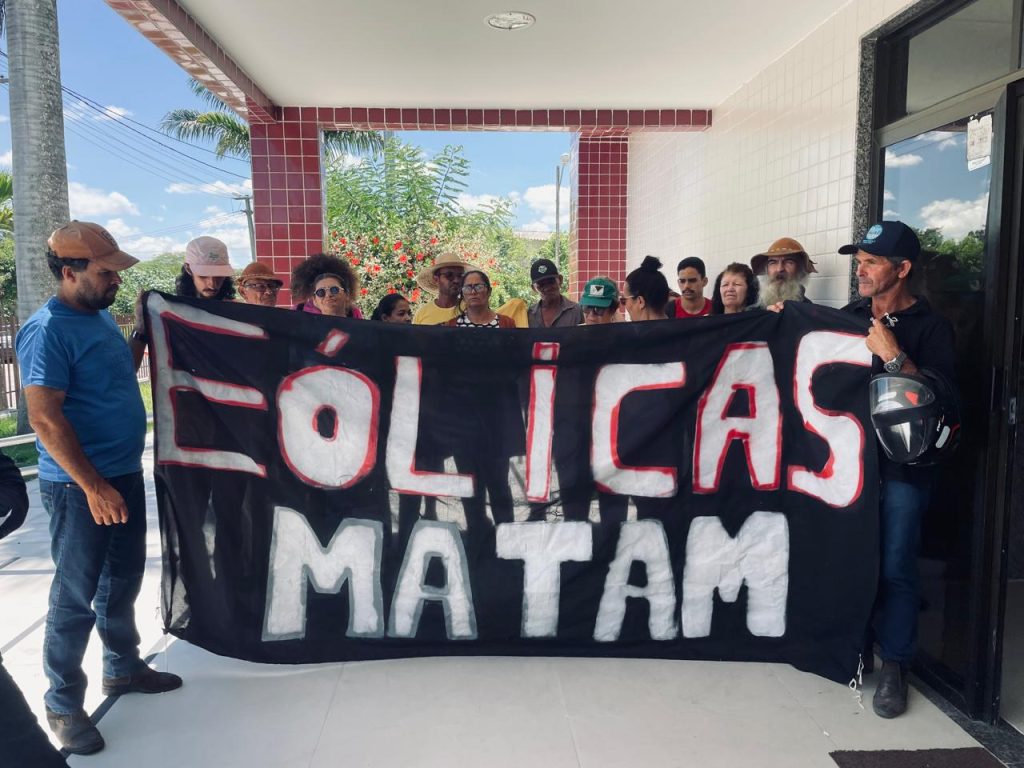 A imagem mostra um grupo de pessoas reunidas em frente a um prédio, segurando um grande banner preto com letras brancas pintadas à mão. O texto no banner diz “EÓLICAS MATAM”, em português, indicando um protesto contra projetos de energia eólica. Ao fundo, há paredes revestidas de azulejos brancos, colunas de tijolos vermelhos, além de plantas e palmeiras que compõem o cenário externo. É uma cena de manifestação pública, marcada pela crítica a empreendimentos ligados às turbinas de vento.