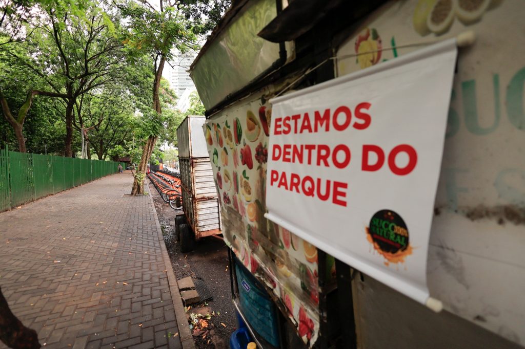 A foto mostra uma calçada de parque com uma cerca verde ao lado e uma fileira de bicicletas laranjas de uso compartilhado. À direita, há uma pequena barraca de venda de sucos naturais, decorada com imagens de frutas e um banner branco com letras vermelhas dizendo “ESTAMOS DENTRO DO PARQUE”. O espaço é arborizado, com árvores altas e caminho pavimentado, transmitindo a ideia de lazer urbano e convivência em área verde.