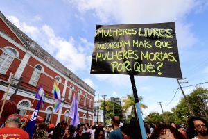 A foto mostra uma manifestação pública em frente a um prédio colonial vermelho e branco. Há várias pessoas reunidas na rua, segurando bandeiras e cartazes. Um dos cartazes em destaque, com letras verdes sobre fundo preto, traz a frase em português: “Mulheres livres incomodam mais que mulheres mortas. Por quê?”.