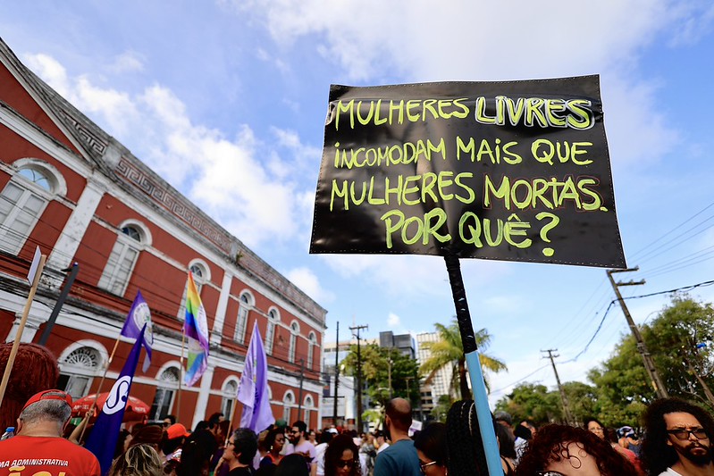 A foto mostra uma manifestação pública em frente a um prédio colonial vermelho e branco. Há várias pessoas reunidas na rua, segurando bandeiras e cartazes. Um dos cartazes em destaque, com letras verdes sobre fundo preto, traz a frase em português: “Mulheres livres incomodam mais que mulheres mortas. Por quê?”.