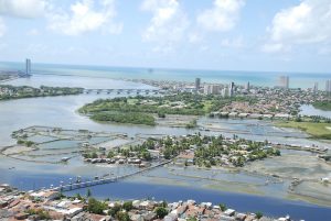 É uma foto aérea que mostra partes do centro e da zona sul do Recife. Em primeiro plano aparecem áreas residenciais cercadas por canais e pequenas ilhas. Uma longa passarela atravessa a água, ligando dois trechos da cidade. Mais ao fundo, há uma ponte maior sobre um corpo d’água e, na linha do horizonte, surgem os prédios altos próximos ao mar. A imagem combina a urbanização com a geografia costeira característica da capital pernambucana.