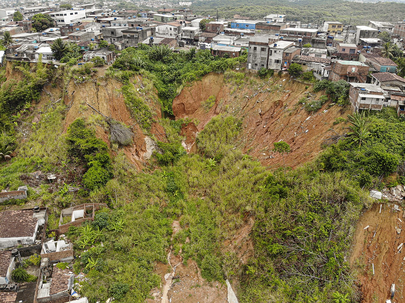 A foto mostra um grande deslizamento de terra em meio a uma área urbana. O solo cedeu e formou uma depressão profunda, com encostas íngremes de terra exposta em tom avermelhado. Há vegetação crescendo de forma irregular nas paredes e no fundo do deslizamento, sinal de recuperação natural. Ao redor, várias casas estão muito próximas da borda, transmitindo a sensação de risco para quem vive ali. É uma cena que evidencia como a instabilidade do solo pode afetar diretamente comunidades e construções.