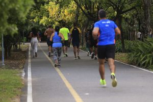 A foto mostra um grupo de pessoas caminhando e correndo em um parque. Elas estão em uma pista pavimentada com linhas amarelas e brancas que delimitam o caminho. Ao redor, há muitas árvores e vegetação, criando um ambiente verde e agradável. As pessoas usam roupas esportivas, como camisetas, shorts e leggings, e parecem estar praticando exercícios ou aproveitando o espaço para lazer.