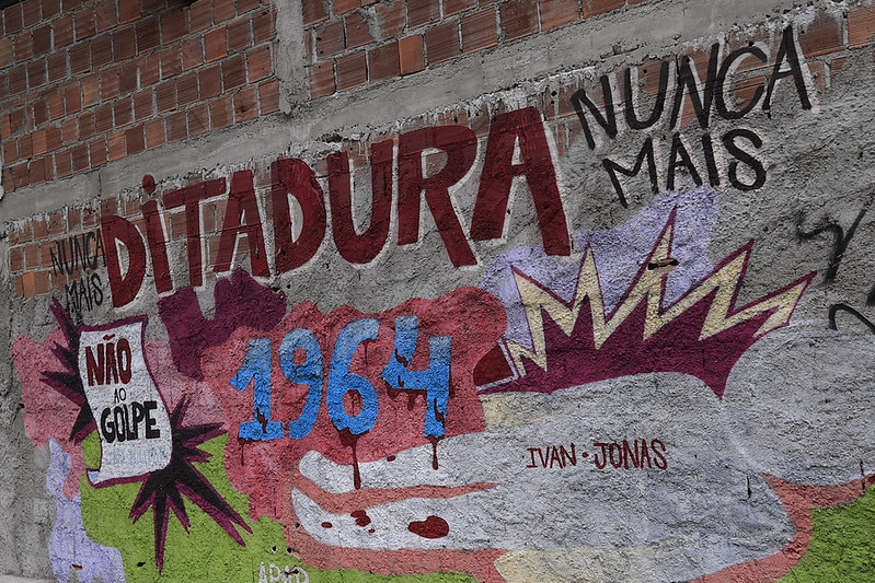 O mural apresenta a frase em destaque “Ditadura Nunca Mais”, pintada em cores fortes como vermelho, azul e amarelo. O número 1964 aparece em azul com tinta escorrida, remetendo ao golpe militar brasileiro. Há também a inscrição “Não ao Golpe” em uma caixa branca com efeito de explosão, compondo uma obra de protesto político e memória histórica.