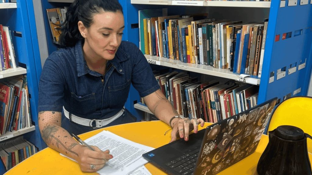 Foto de Milena Barros Gomes, mulher de pele clara, com cabelos pretos presos em um rabo de cavalo. Ela veste um macacão jeans azul com cinto branco e tem tatuagens nos braços. Está sentada a uma mesa amarela, escrevendo. À sua frente, há um computador e, ao fundo, estantes azuis repletas de livros coloridos.