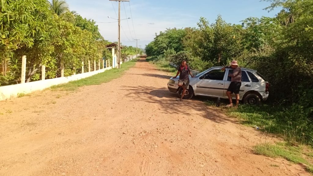 Em uma estrada de terra no campo, cercada por vegetação e uma cerca de madeira à esquerda, há um carro prateado parado parcialmente sobre a grama à direita. Duas pessoas estão próximas: uma em pé diante do veículo e outra apoiada na porta. Ao fundo, aparecem árvores, arbustos, alguns postes de energia e o céu claro.