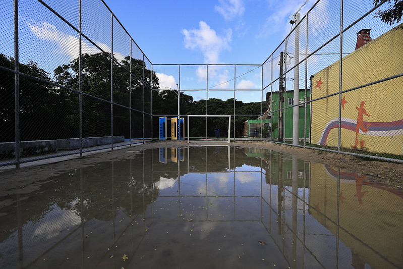 A foto mostra uma quadra esportiva cercada, provavelmente de futebol ou futsal, parcialmente coberta por uma grande poça d’água que reflete o céu, o alambrado e as construções ao redor. Ao fundo, aparecem dois brinquedos de cimento para crianças escalarem. À direita, há uma parede amarela com um mural colorido de um jogador de futebol estilizado, estrelas e linhas curvas em vermelho e azul. O reflexo da cena na água cria um efeito visual simétrico e marcante.
