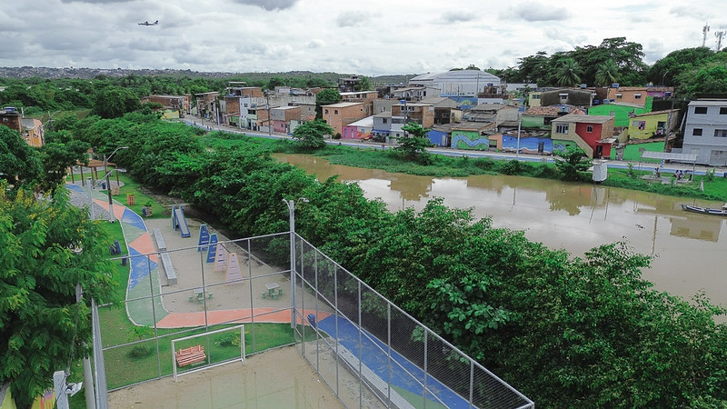 A foto mostra uma vista aérea de um bairro cortado por um rio de águas barrentas. De um lado, há um parque com quadra esportiva cercada, caminhos para caminhada, bancos e equipamentos de exercícios ao ar livre. Do outro lado, vê-se uma fileira de casas coloridas em tons de marrom, vermelho, azul e verde. As margens do rio são cobertas por vegetação densa e, ao fundo, um avião aparece no céu, sugerindo a proximidade de um aeroporto.