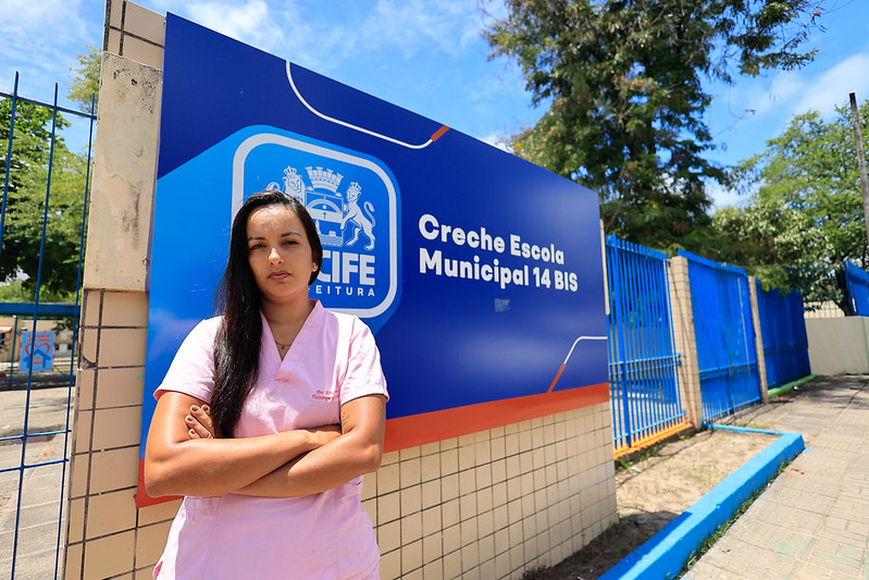 Na foto vemos Aline Torres, em frente ao portão de uma creche municipal em Recife. Ela veste um uniforme rosa claro e está de braços cruzados, transmitindo uma postura firme e confiante. Aline tem pele clara, cabelos escuros presos para trás e aparece em pé diante de um grande painel azul e laranja com o brasão da prefeitura. O cenário ao redor inclui uma parede revestida de azulejos, árvores ao fundo e um caminho pavimentado.