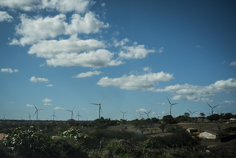 A foto mostra um cenário rural com colinas suaves, onde várias turbinas eólicas brancas se erguem contra o céu azul com nuvens espalhadas. No primeiro plano, há vegetação baixa e algumas construções pequenas, como uma casa ou galpão. É uma paisagem que combina elementos naturais com tecnologia de energia renovável.