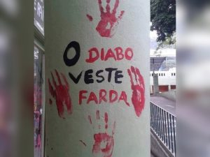 A imagem mostra um pilar de concreto cilíndrico em espaço público. Sobre ele, há várias marcas de mãos pintadas em vermelho, como se fossem impressões deixadas com tinta. Entre essas marcas, aparece uma frase escrita em português: “O diabo veste farda”, em letras pretas e vermelhas. O conjunto transmite uma mensagem de protesto ou crítica, reforçada pelo contraste entre as cores e pela repetição das mãos. Ao fundo, vê-se parte de uma calçada com corrimão e alguns prédios.